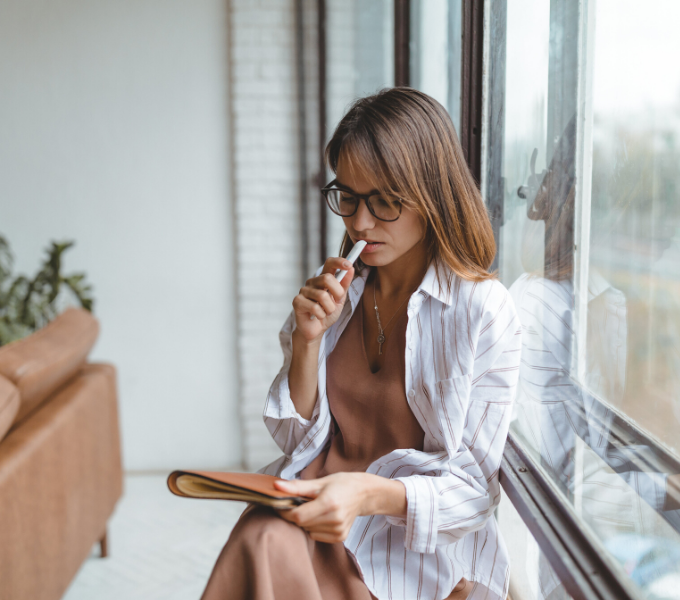 Leadership and executive coaching - Newsky Consulting - A brunette lady sitting by a window with a notebook, she looks like she's thinking